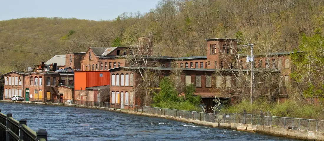 Old red brick mill buildings along a river below a wooded hillside