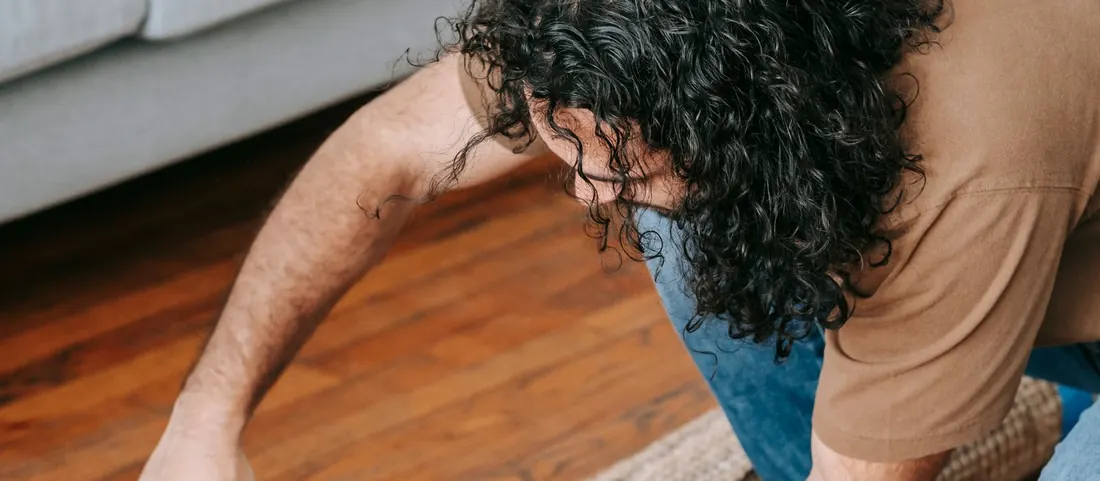 Man taping a rolled carpet closed on a wood floor beside a grey sofa