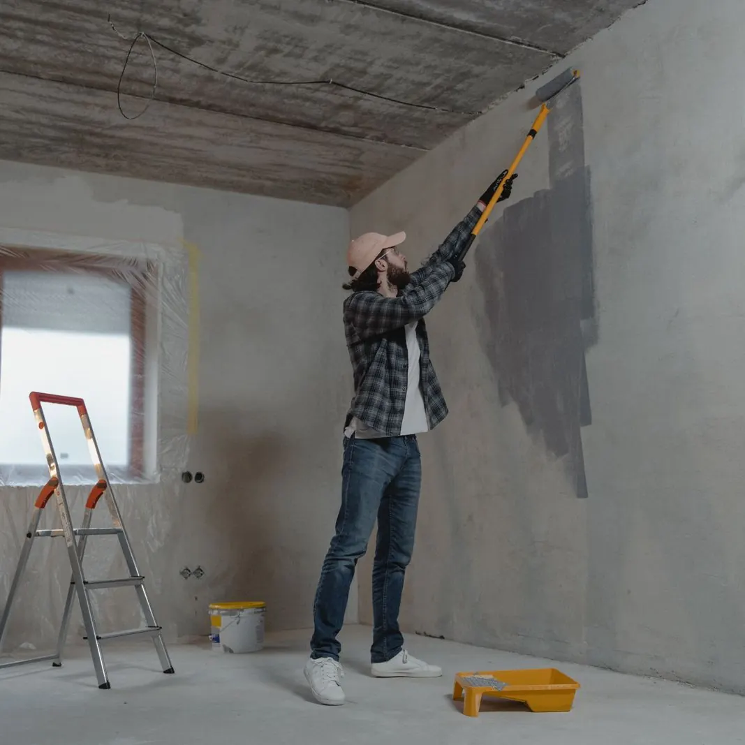 Painter rolling grey paint onto a bare wall beside a step ladder and paint tray